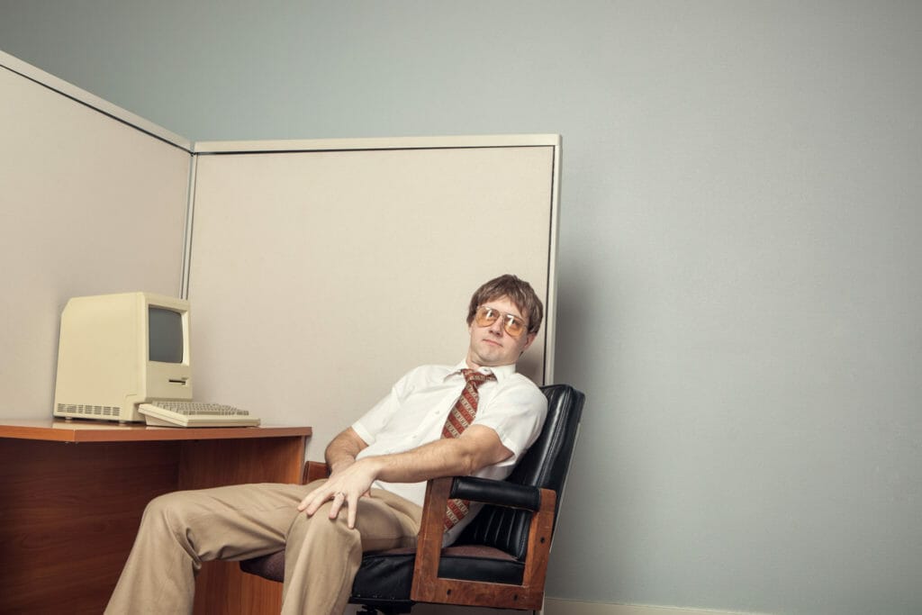 A lazy I.T. computer technician leans back in his chair in his office cubicle sitting in front of his desk with a vintage 1980s computer.  Horizontal with copy space.